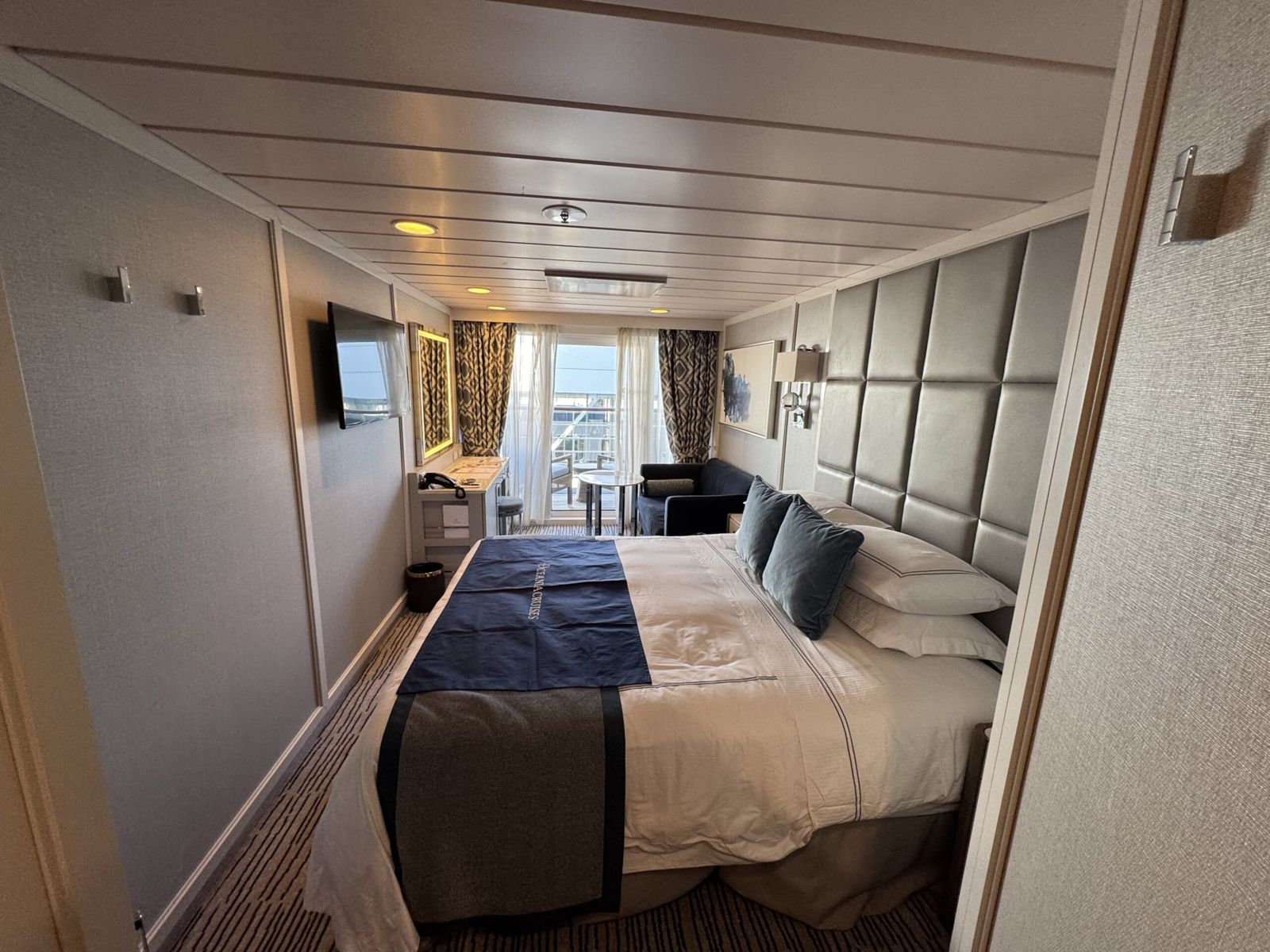 Our room on the Insignia. The walls are a soft brown. The bed dominates the room in the foreground. White sheets and blue pillows and topper. A tv is on the wall, a desk and lounge are in the far end, where doors open to a balcony with two seats and a small table. 