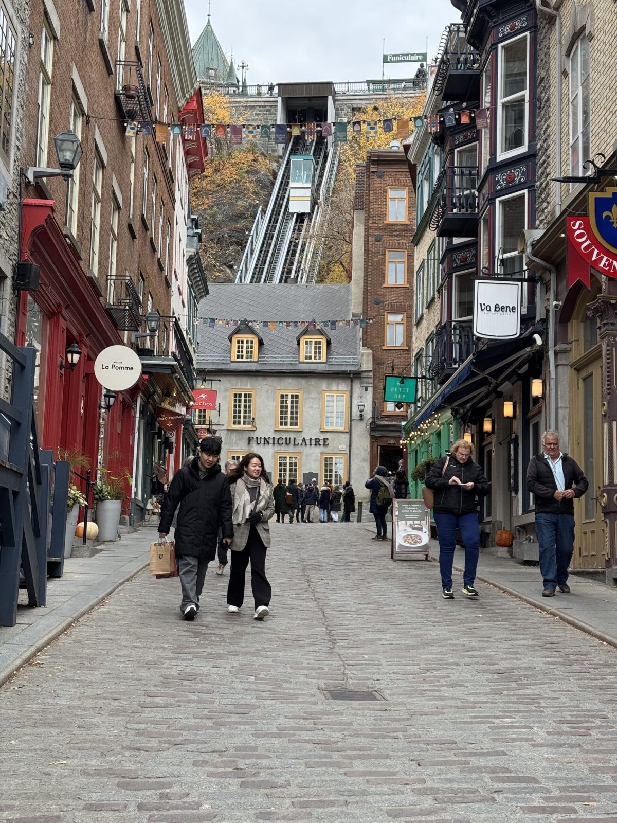 A quaint street in Quebec City. Each side of the street has colourful store fronts. There are people walking up and down the street. In the distance a building sits in front of a steep hill up the you can see a vernacular. A type of train that takes people up the hill in stead of steps. 