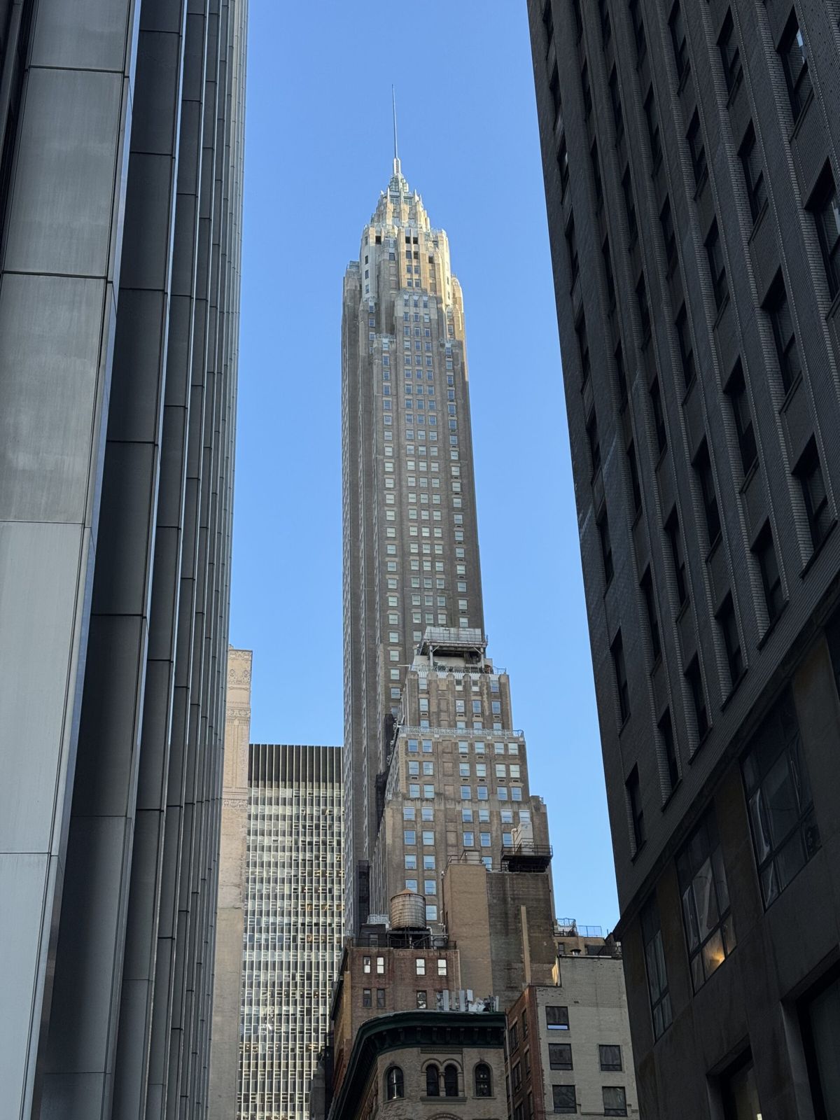 &quot;A tall, historic skyscraper known as the Woolworth Building, featuring a Gothic-style architecture with ornate detailing and a greenish, copper roof against a clear blue sky. The building's façade is decorated with intricate terracotta tiles, and its towering spire rises prominently in the Manhattan skyline.&quot; 