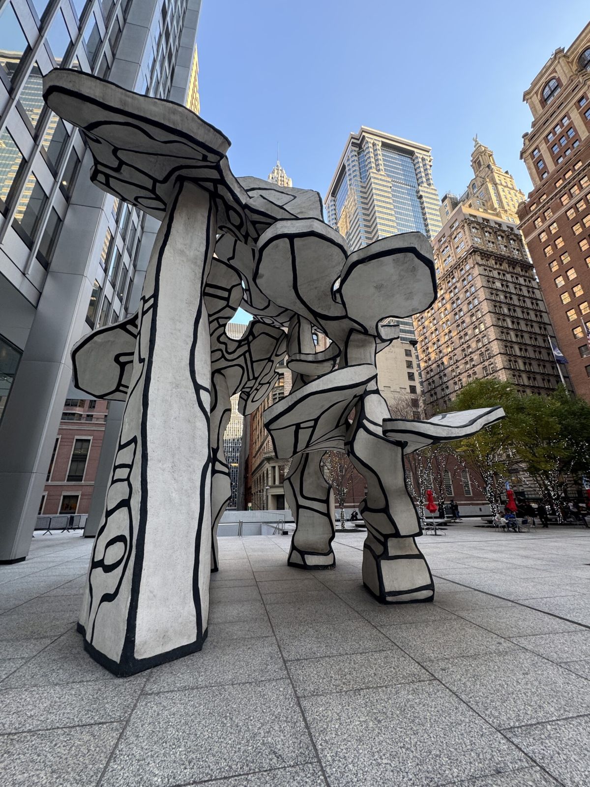 The a large abstract sculpture situated in an urban setting, surrounded by tall buildings. The sculpture consists of multiple interconnected, irregularly shaped forms with black outlines, reminiscent of a drawing brought to life. The ground is paved with light gray tiles, and a few people can be seen walking around, adding a sense of scale. The scene captures a modern, architectural aesthetic, with the city skyscrapers towering in the background under a clear blue sky.