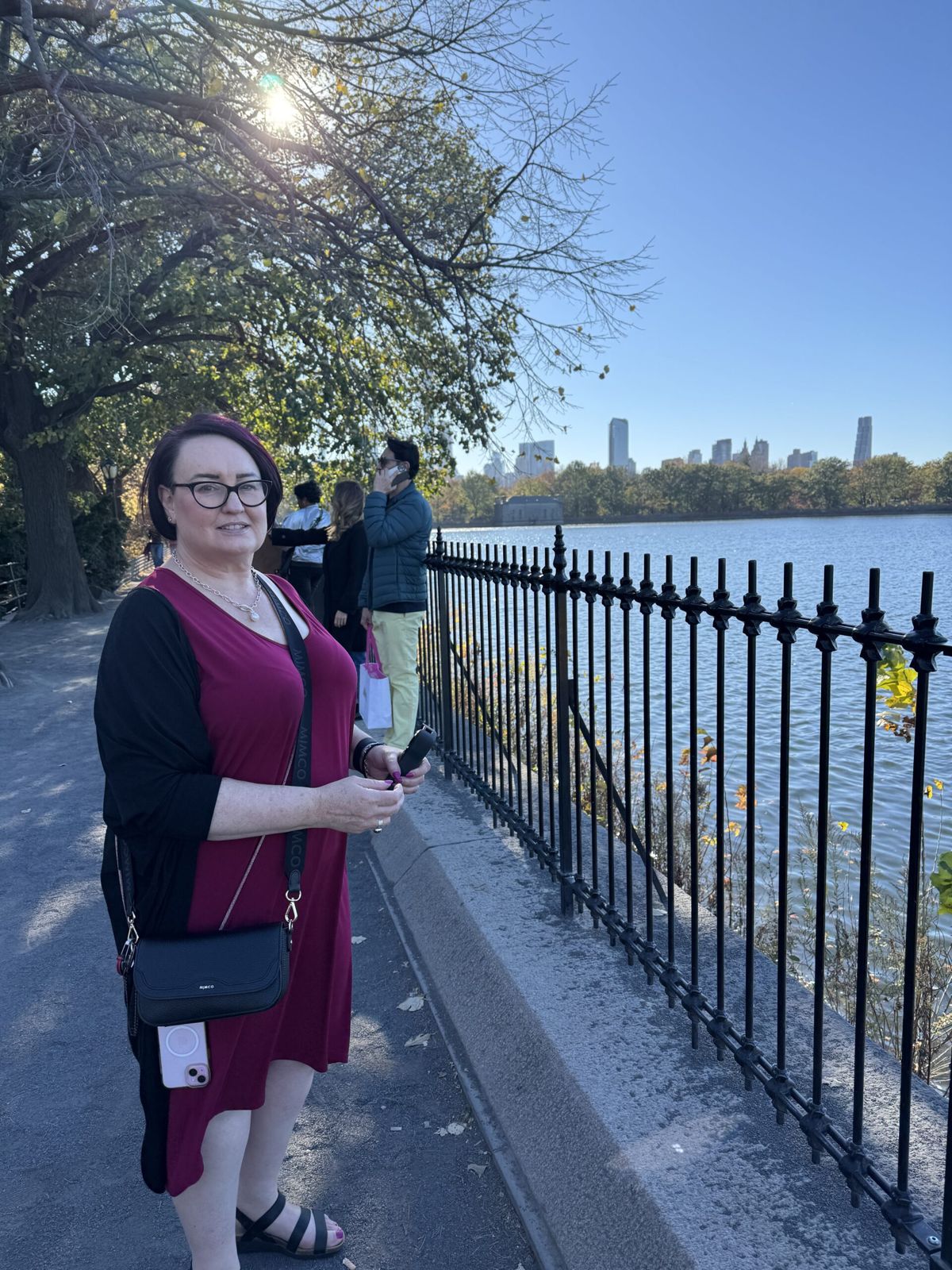 Mardi in front of the reservoir, a fence on her right. She’s wearing a red dress and black cardigan. You can see water and in the distance the skyline of Manhattans buildings along 57th street.