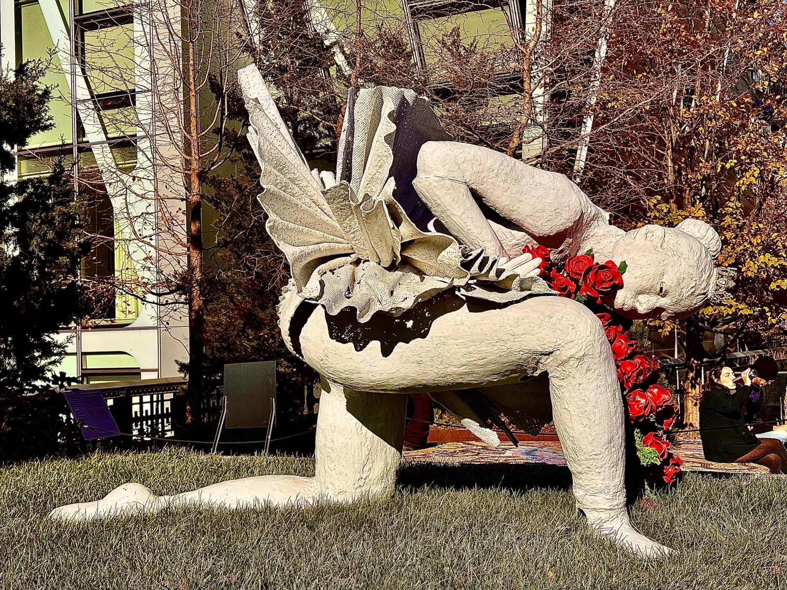 A white statue of a ballerina. She’s leaning forward on one knee. She’s holding a red rose. 