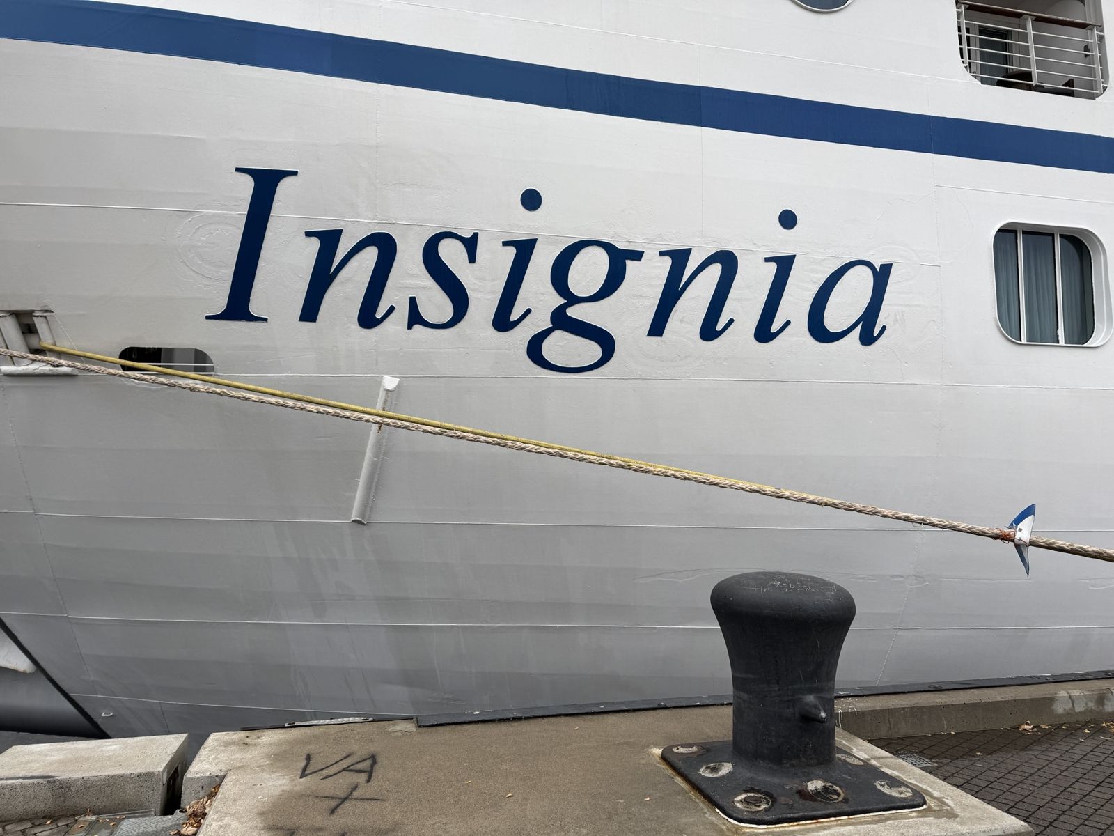 The side of our ship. Showing its name insignia and ropes tying it to the dock. The ship is white and blue lettering. 